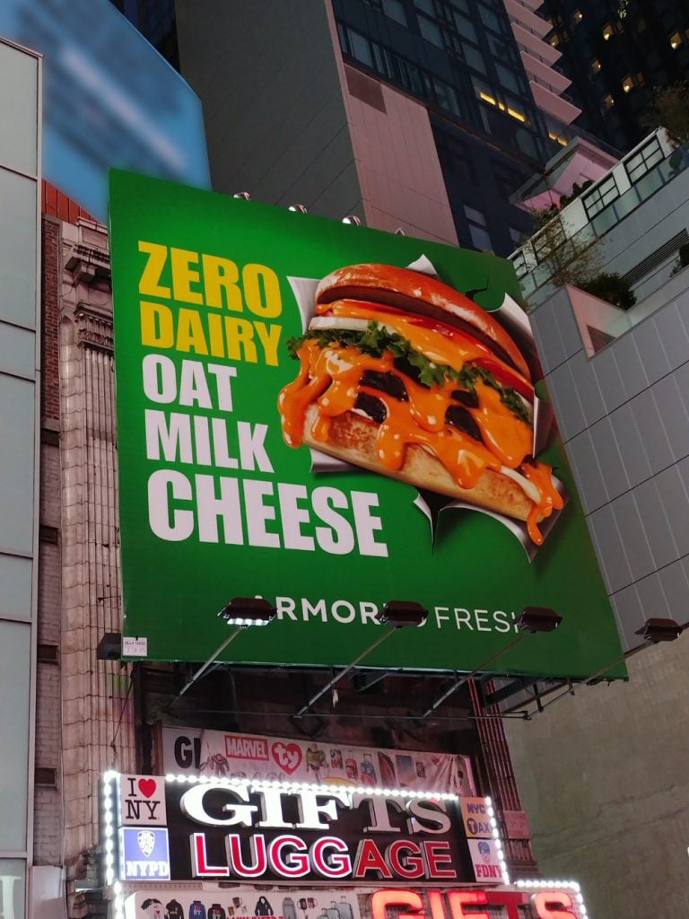 Times square billboard by vegan cheese brand Armored Fresh. A large vertically oriented static board with a green background and a burger with melty cheese creating a 3D effect. The text reads ZERO DAIRY OAT MILK CHEESE.