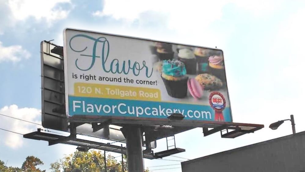 Billboard ad by a cupcake shop with a large image of cupcakes and other desserts next to the text FLAVOR IS RIGHT AROUND THE CORNER! The business address and website are below.