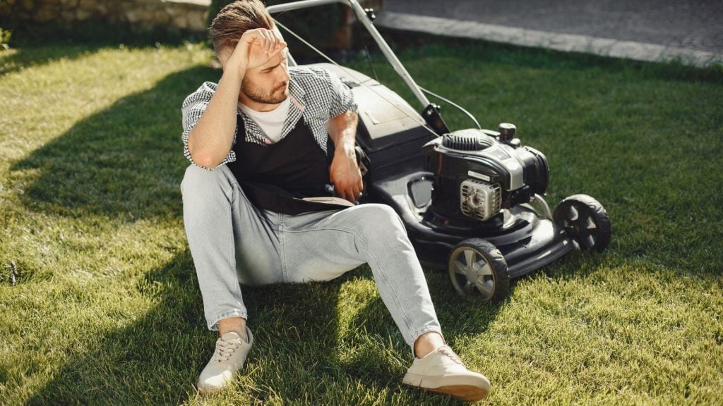 A picture of a man sitting next to a lawnmower exhibiting signs of stress.