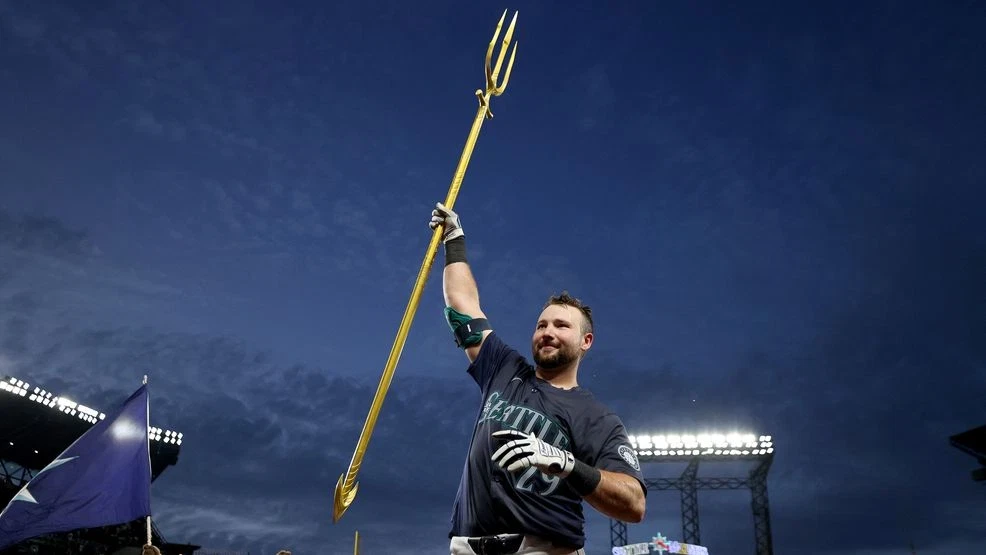 A photo of Seattle Mariner's baseball player Cal Raleigh holding up the Mariner's trident  with the stadium lights in the background and a cloudy evening sky.