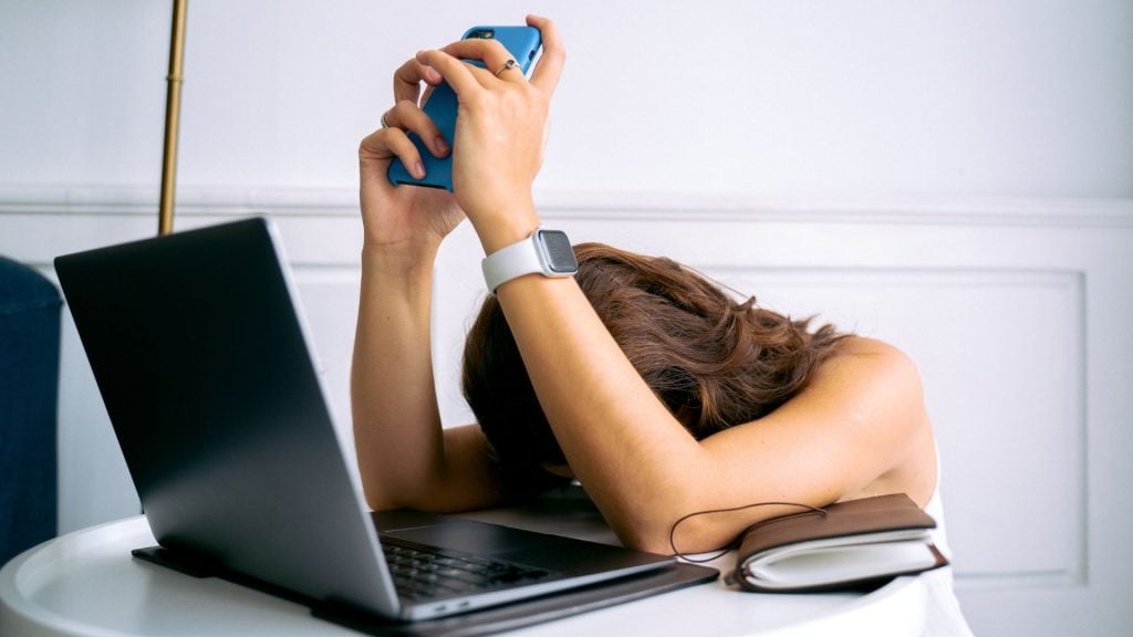 A photo of a women exhibiting digital fatigue at her computer holding her phone with her head resting defeatedly in between her arms slouched at a table.
