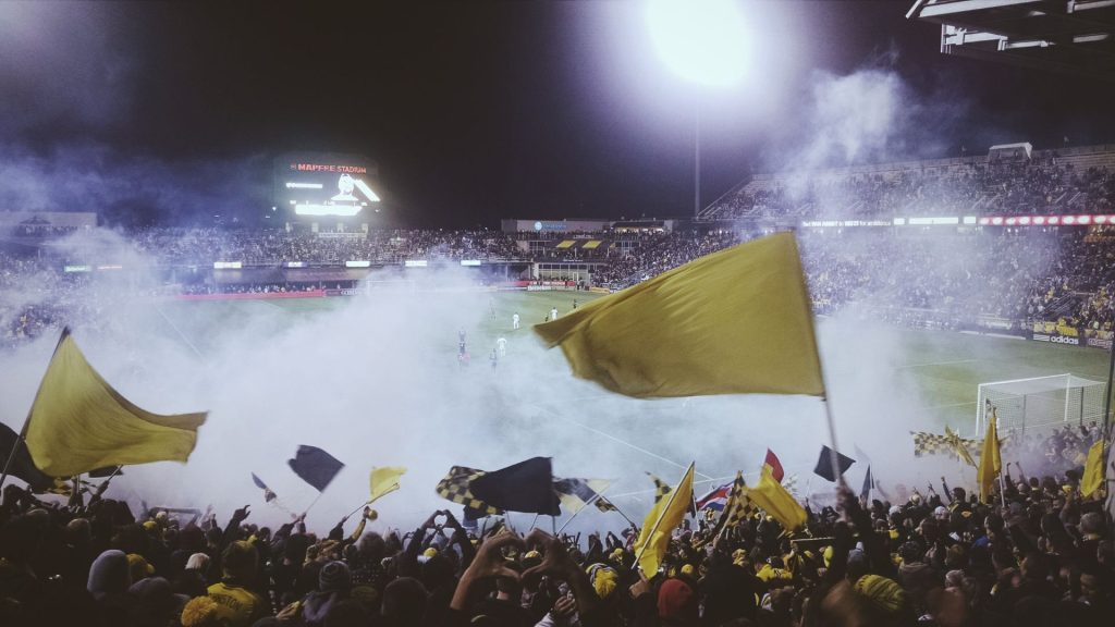 A stadium of fans celebrating waiving flags with smoke in the stands at a soccer match.