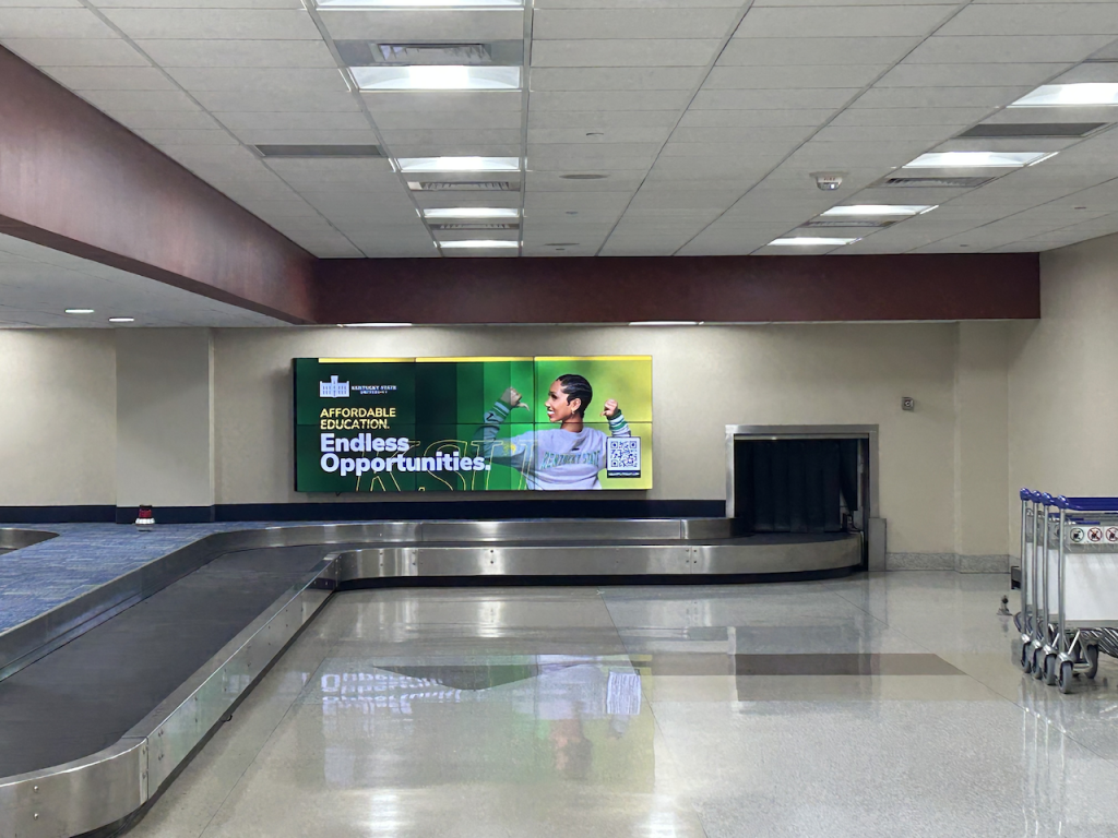 A digital airport ad above a baggage claim for Kentucky State University.