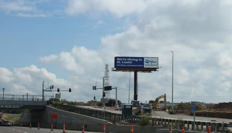 Roadside billboard by Gateway Arch area in St. Louis displaying a hiring message above a highway interchange with traffic lights, construction equipment, and urban infrastructure in the background.