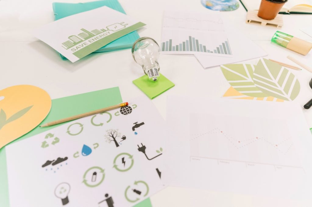 Sustainable business workspace featuring green marketing materials, energy-saving infographics, data charts, a lightbulb, and eco-friendly icons spread across a white desk.