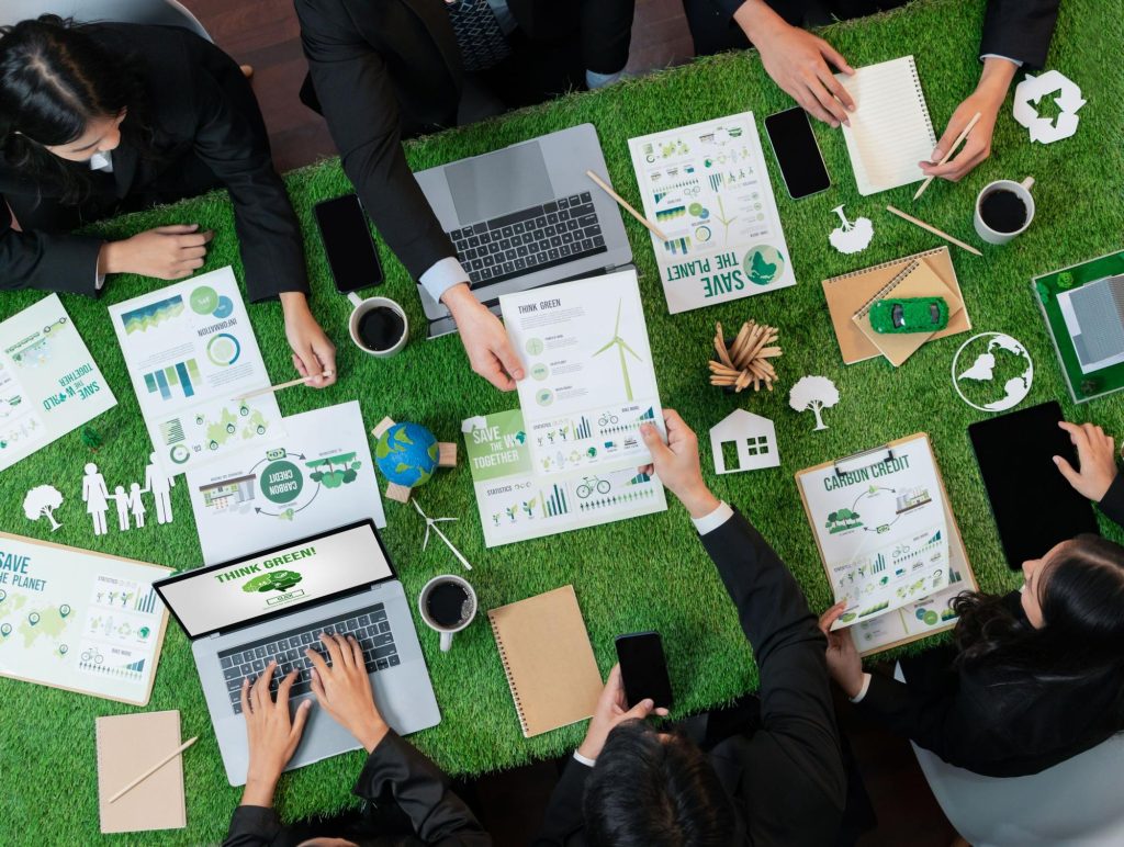 Top-down view of a business team engaged in green marketing strategy, working around a grass-covered table with eco-friendly infographics, laptops, and carbon credit documents.
