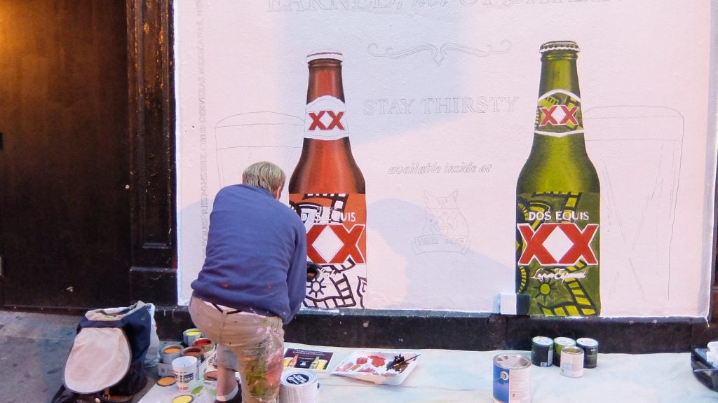 Artist painting a large branded wall mural advertisement featuring beverage bottles, illustrating one of the creative types of outdoor advertising that uses hand-painted street art to attract attention in urban environments.