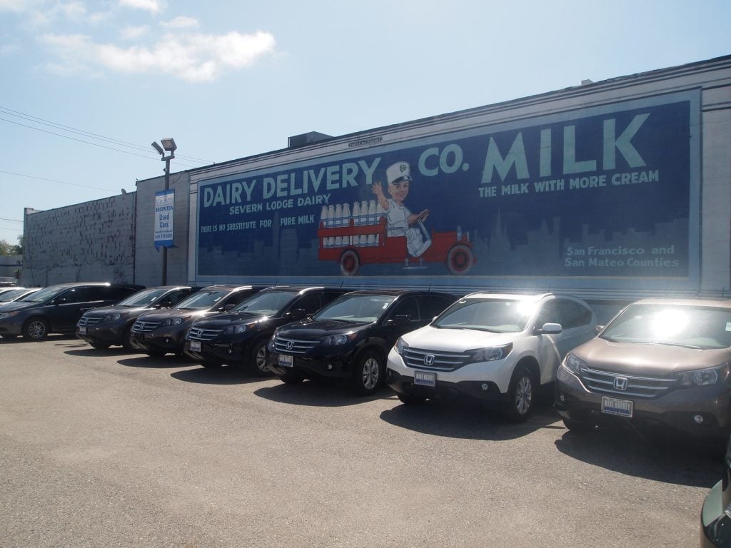 Large wall mural advertisement painted on the side of a building above a car lot, demonstrating one of the classic types of outdoor advertising that uses long-term, street-level visibility to reach local audiences.