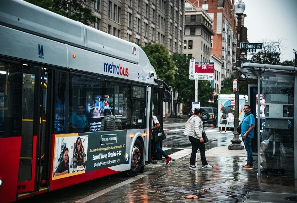Urban street scene with a city bus displaying a large side-panel advertisement at a bus stop, illustrating transit advertising as one of the prominent types of outdoor advertising used to reach commuters and pedestrians in busy city environments.