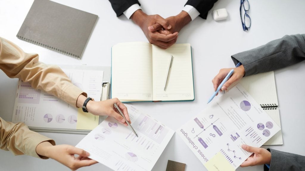 Overhead view of a business team's hands pointing at diverse charts and a note labeled "Business challenges" on a desk, illustrating a key step in how to conduct market research.