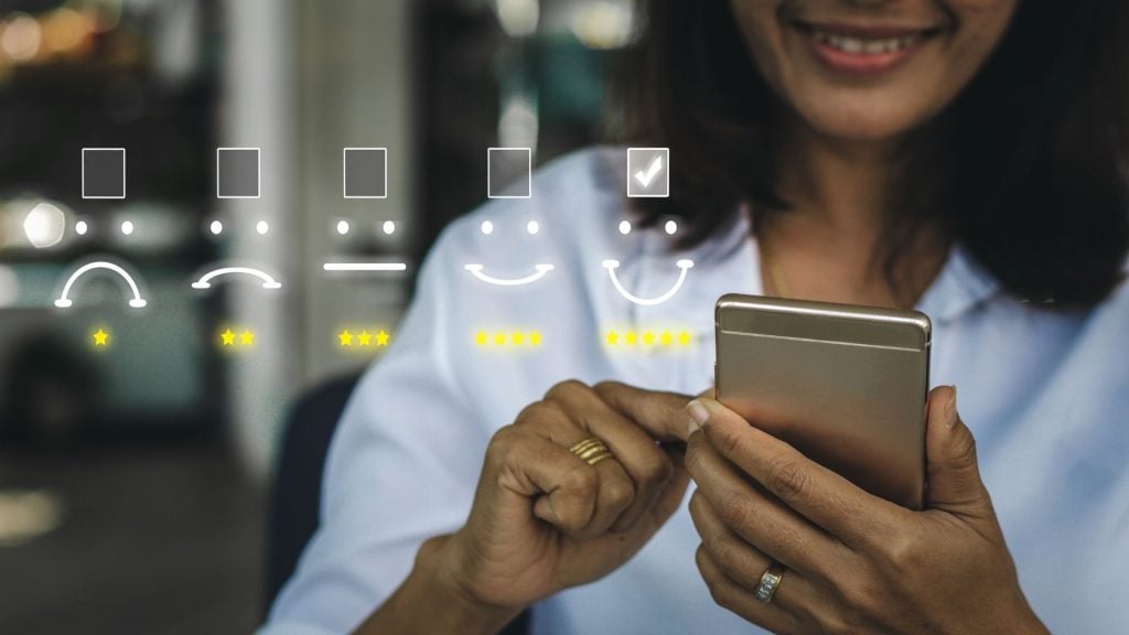 A smiling woman holds a smartphone displaying five graphic customer service rating options, from sad to happy, as part of her process on how to conduct market research.