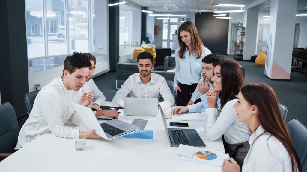 A collaborative business team sits around a conference table with laptops and a detailed pie chart infographic, working together on a project that demonstrates how to conduct market research.