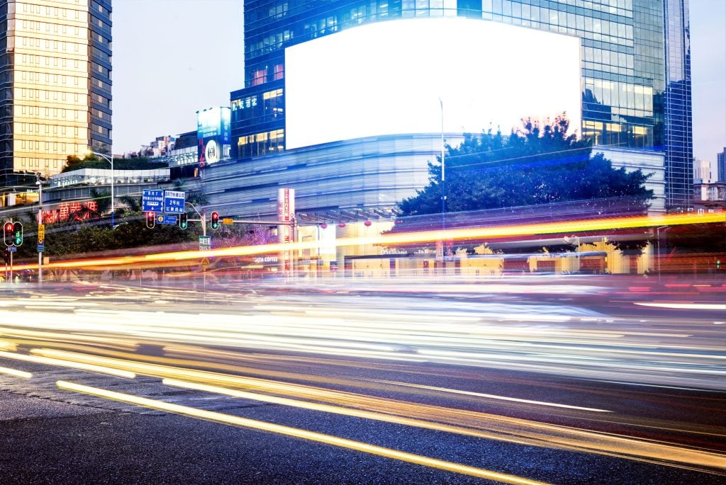 A dynamic city street scene at night with light trails and a prominent blank white billboard, illustrating the high-visibility canvas available for those researching how to incorporate ooh advertising digital campaign placements in urban centers.
