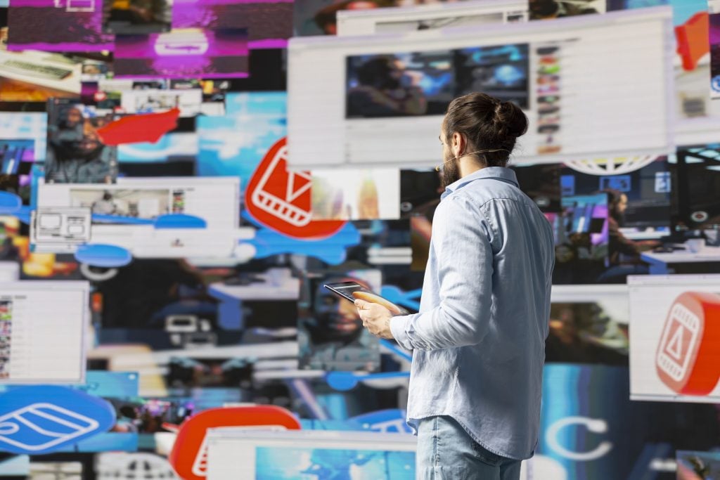 A professional presenter holding a tablet in front of a dynamic wall of digital screens displaying diverse media content and icons, serving as a conceptual representation for how to incorporate ooh advertising digital campaign strategies in a complex, multi-screen digital landscape.