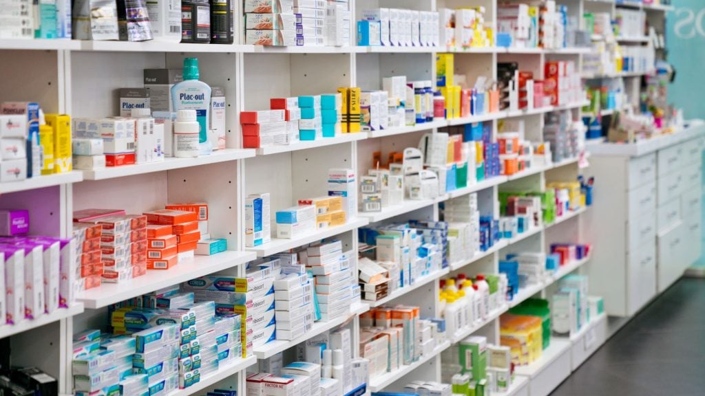 Wide-angle view of organized white pharmacy shelves stocked with various over-the-counter medications and health products, used for pharmacy advertising retail displays.