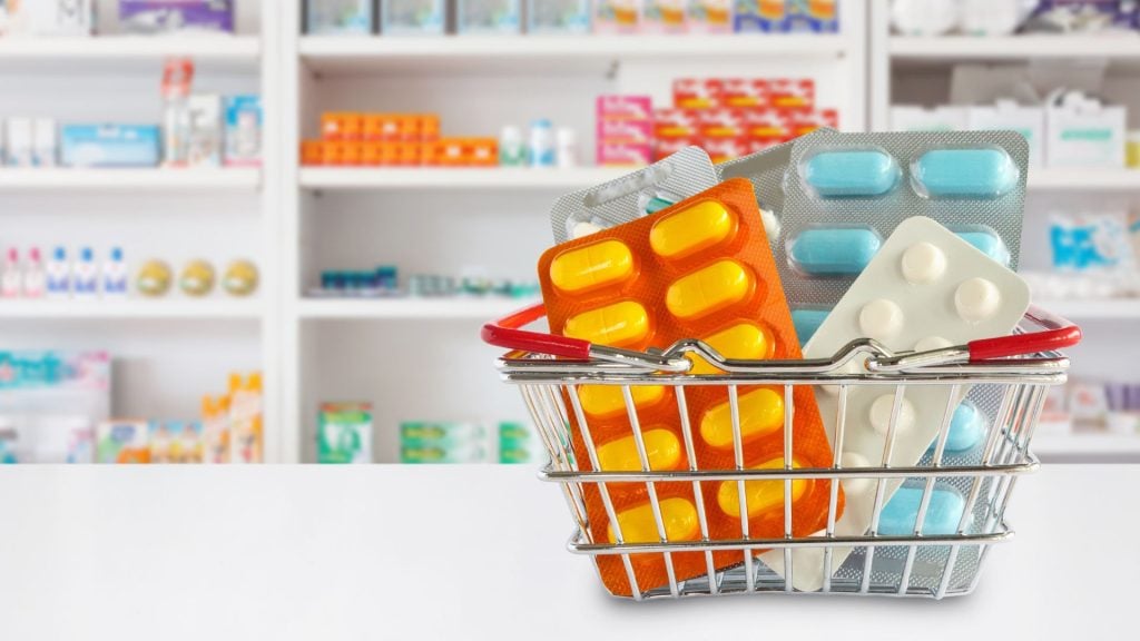 A small metal shopping basket filled with orange and blue pill blister packs on a white counter, illustrating consumer-focused pharmacy advertising in a retail setting.