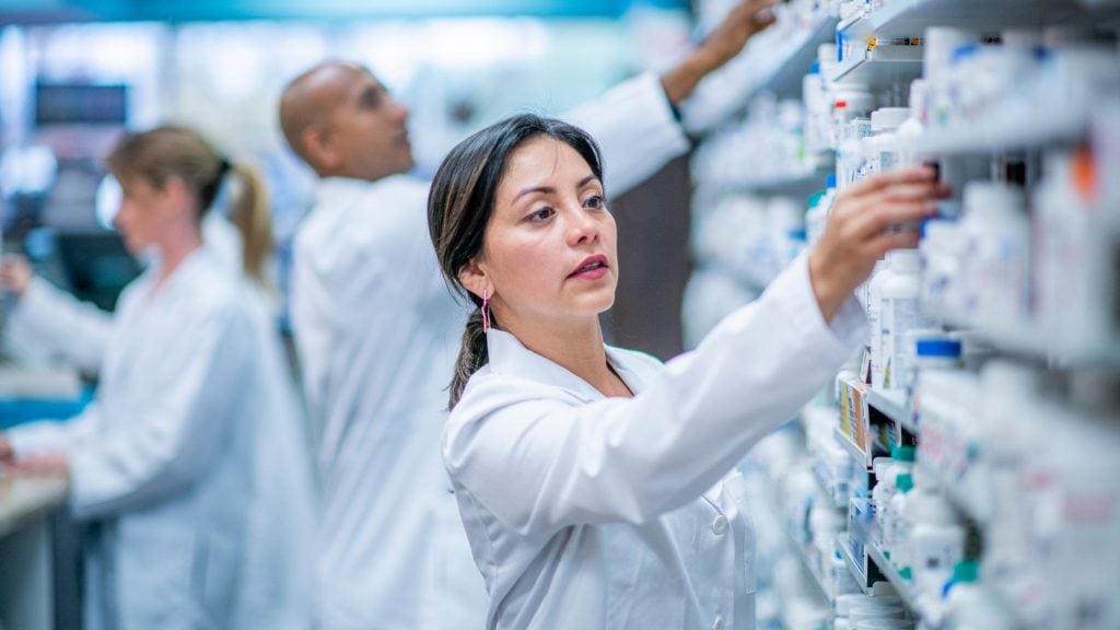 A professional female pharmacist in a white lab coat organizing medicine bottles on a shelf, representing the trust and expertise highlighted in pharmacy advertising.
